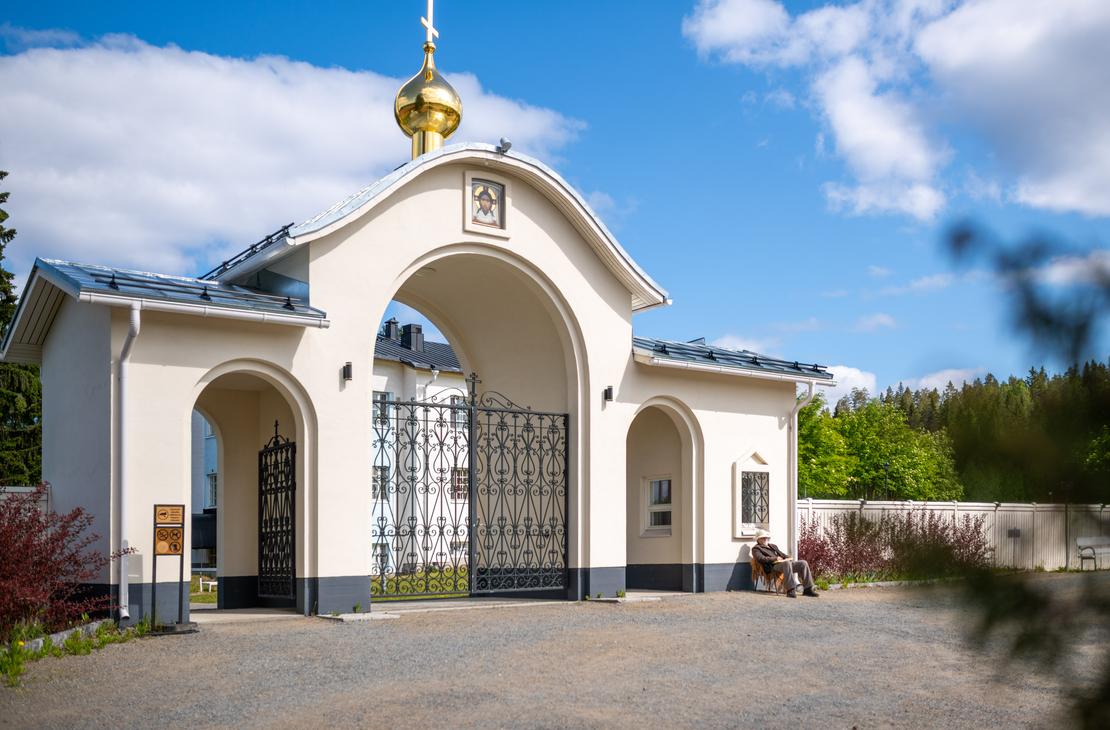 Valamon luostarin vaalea, kaari-aukkoinen porttirakennus, jonka yläpuolella on kultainen sipulikupoli ja risti. Koristeellinen, musta metalliportti on suljettu. Oikealla rakennuksen seinustalla istuu henkilö penkillä aurinkoisena päivänä.