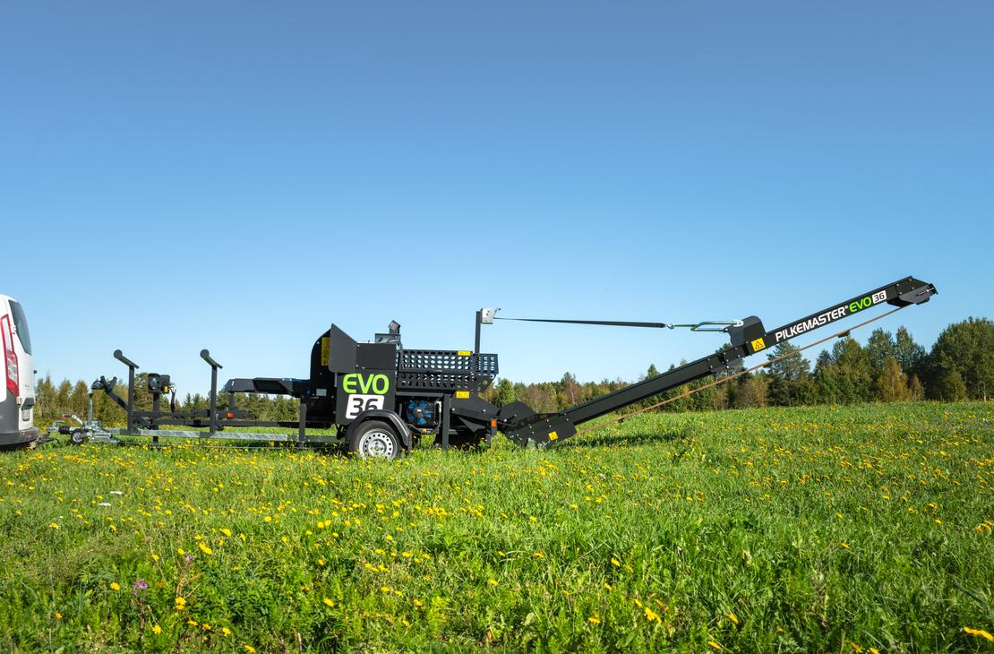 KLAPikosen musta ja vihreä Pilkemaster EVO 36 -klapikone seisoo aurinkoisella, keltaisten voikukkien täplittämällä niityllä valkoisen pakettiauton perässä, kirkkaansinisen taivaan alla.