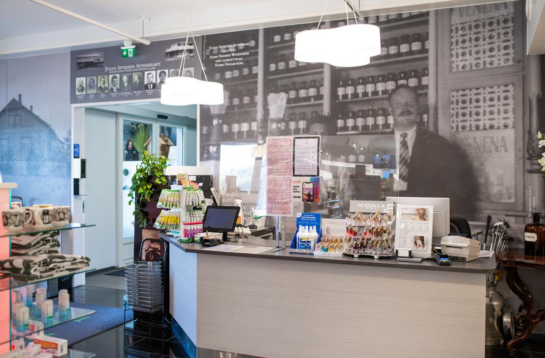 Kuva esittää Juuan Apteekin sisätilaa ja palvelutiskiä. Etualalla on harmaa palvelutiski, jonka takana on seinällä mustavalkoinen, suuri valokuva vanhanaikaisesta apteekista tai apteekkarista hyllyineen. Tiskillä on erilaisia apteekkituotteita. Tila on hyvin valaistu.