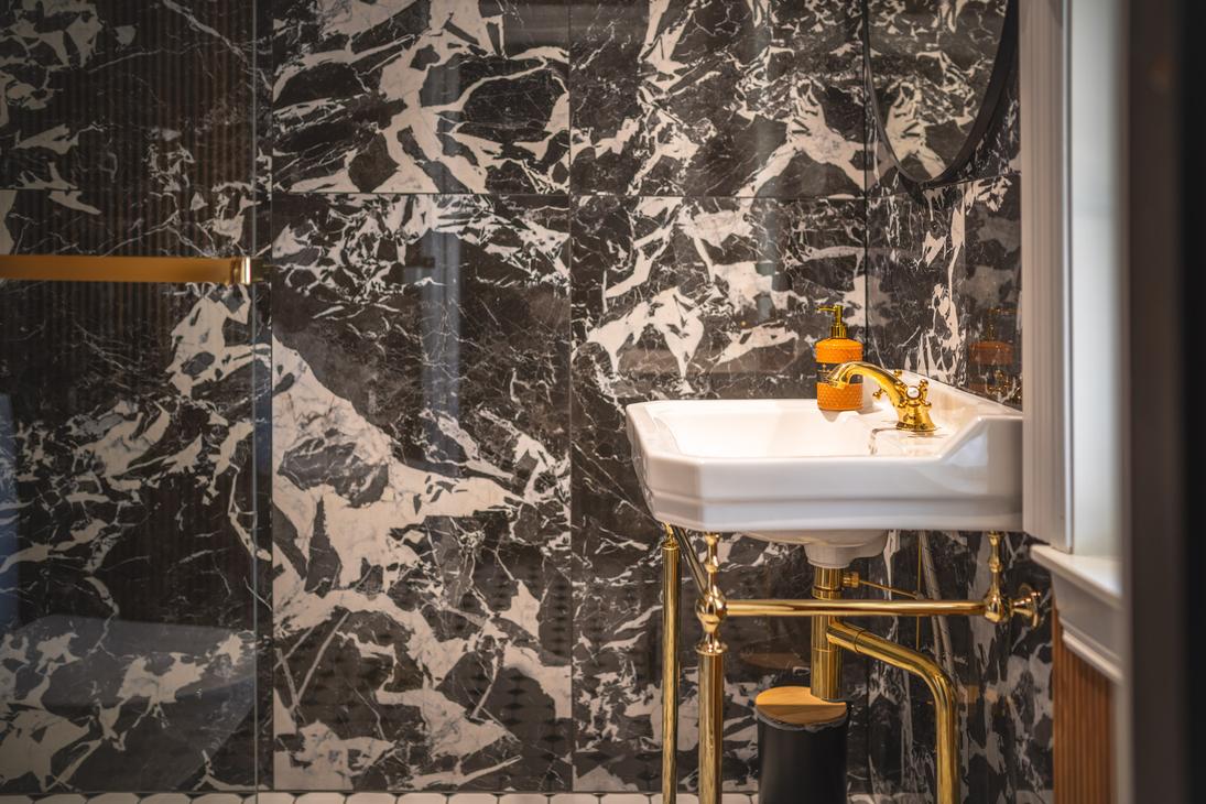 Kuva esittää Matkailukeskus Puu-Juuka Oy:n kylpyhuonetta tai wc-tilaa. Seinät on kaakeloitu dramaattisella, tummalla marmorikuviolla (mustaa ja valkoista). Etualalla on valkoinen, perinteinen pesuallas (lavuaari), joka on asennettu kiiltäviin, kultaisenvärisiin metallijalkoihin ja putkiin. Pesualtaan päällä on oranssi kukka tai koriste-esine.