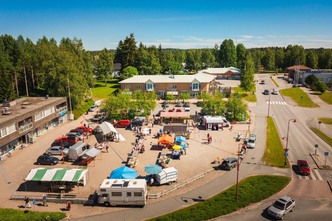 Yläviistosta kuvattu näkymä aurinkoiselta torilta, jossa on useita erivärisiä myyntitelttoja, ihmisiä ja torimyyjiä. Ilomantsin torin laidalla on pysäköitynä matkailuauto ja useita henkilöautoja, ja taustalla siintää S-Marketin liikerakennus sekä metsää.