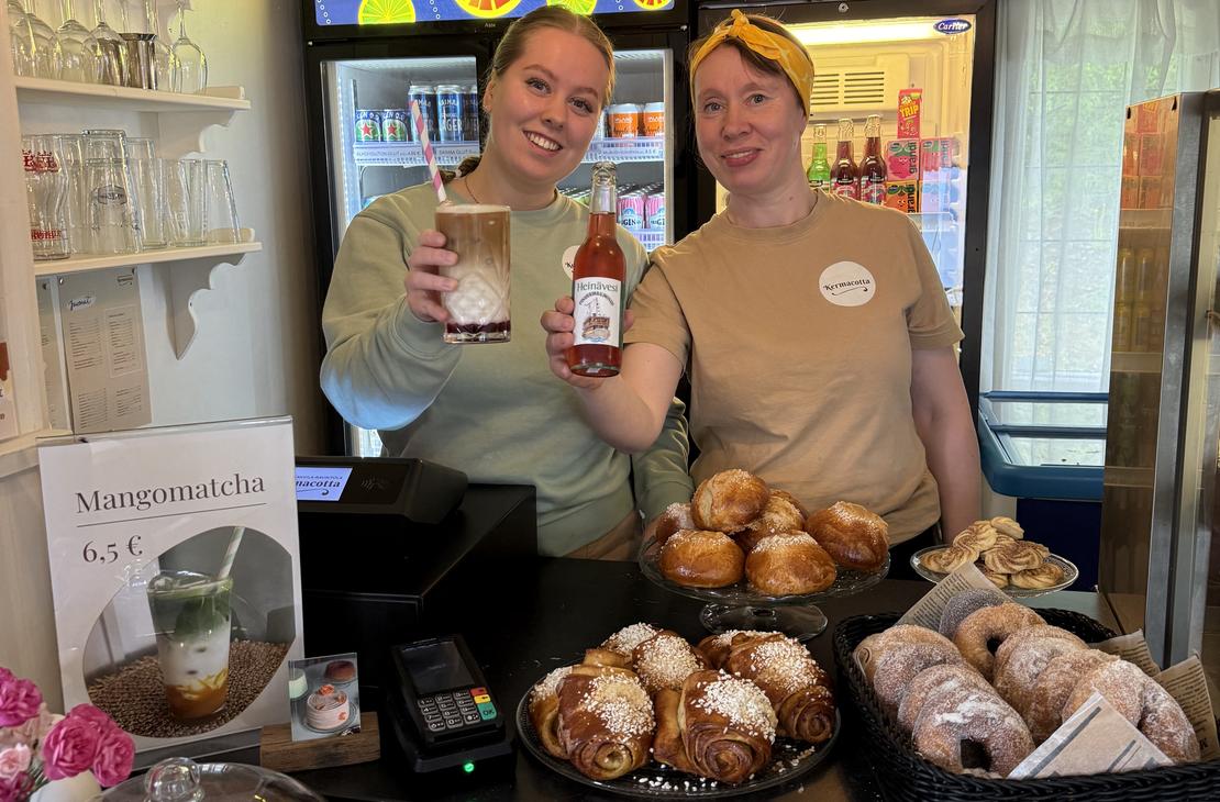 Kaksi hymyilevää työntekijää Satamakahvila-ravintola Kermacotan tiskillä. Toisella työntekijällä on käsissään pullollinen juomaa ja toisella täytetty lasi. Tiskin päällä on esillä houkuttelevia, vastaleivottuja korvapuusteja ja muita leivonnaisia koreissa. Taustalla näkyy kylmäkaappeja juomille.