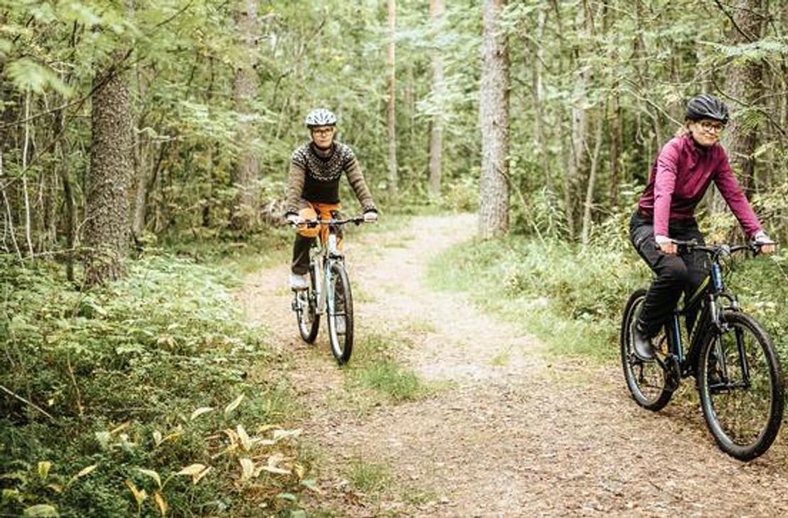 Kaksi henkilöä, nainen etualalla ja toinen henkilö taustalla, pyöräilevät mutkittelevaa hiekkatietä pitkin tiheässä metsässä. Nainen etualalla on pukeutunut pinkkiin paitaan ja kypärään, ja hän ajaa tummaa maastopyörää. Taustalla oleva pyöräilijä on pukeutunut tummiin vaatteisiin ja ajaa vaaleampaa pyörää. Polkua reunustaa runsas, vihreä aluskasvillisuus.
