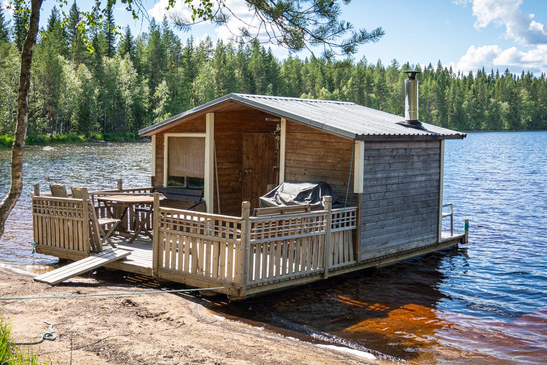 Puurakenteinen saunalautta ankkuroituna hiekkaiselle rannalle tyynen järven äärellä. Lautalla on terassi, jossa on pöytäryhmä ja grilli, sekä korkea metallinen piippu.