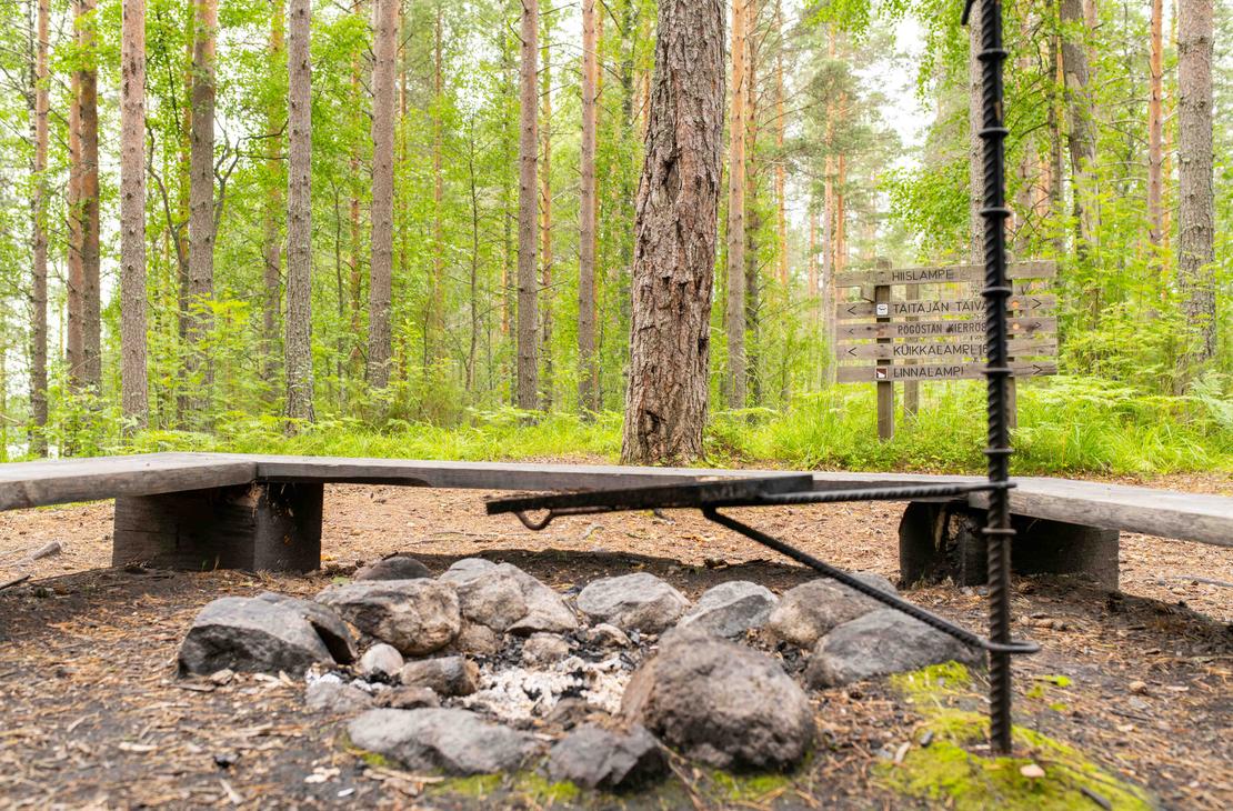 Lähikuva maastossa olevasta nuotiopaikasta ja grilliritilästä. Taustalla näkyy puinen opasteviitta, jossa on reittien ja lampien nimiä, kuten Hiislampi ja Taitajan taival.