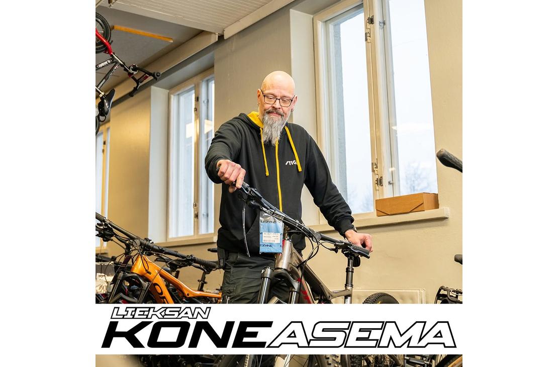 Silmälasipäinen, parrakas mies tarkastelemassa polkupyörää myymälässä. Kuvan alalaidassa on Lieksan Koneasema -tekstilogo.