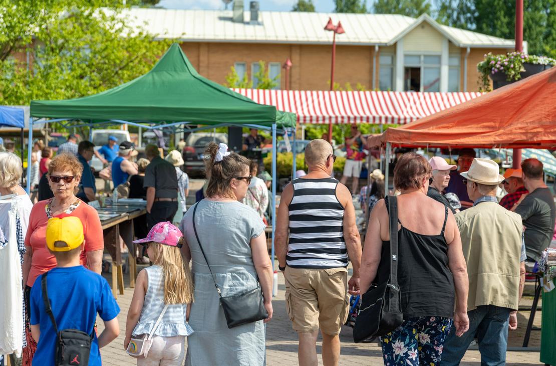Ryhm&auml; kes&auml;asuisia ihmisi&auml; k&auml;velem&auml;ss&auml; Ilomantsissa ulkoilmatorilla vihre&auml;n ja oranssin myyntiteltan v&auml;liss&auml; kohti taustalla n&auml;kyv&auml;&auml; esiintymislavaa, jonka yll&auml; on puna-valkoraitainen katos.