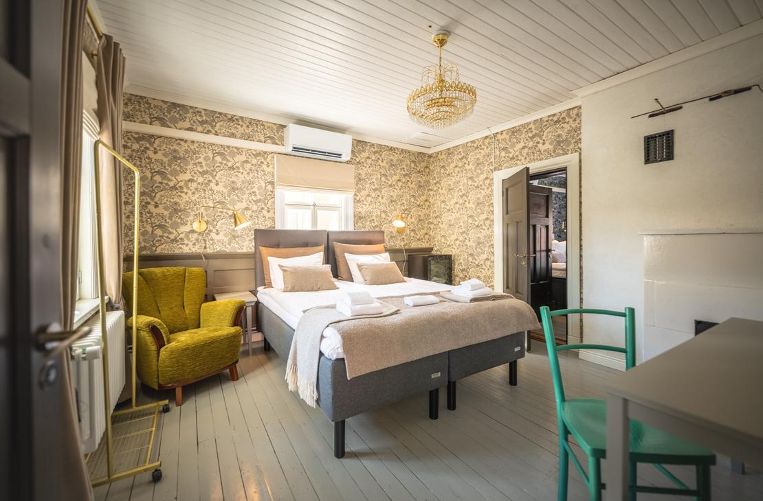 Kuva esittää Matkailukeskus Puu-Juuka Oy:n hotellihuonetta tai majoitustilaa. Huoneessa on leveä, kaksiosainen parisänky neutraalin sävyisillä petivaatteilla. Sängyn takana seinällä on karkea, kivinen tai puinen tapetti. Huoneessa on vaaleansininen puulattia, ja vasemmalla on sammaleenvihreä nojatuoli. Oikealla on pieni pöytä ja kaksi turkoosinsinistä tuolia. Huone on valoisa ja siinä on moderni sisustus.