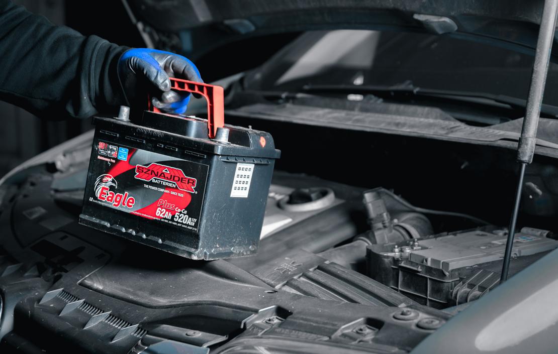 Lähikuva auton akun vaihdosta tai asennuksesta. Kuvassa nähdään käsi sinisessä hansikkaassa nostamassa tai laskemassa uutta, mustaa auton akkua, jossa on punainen kantokahva ja etiketti, harmaasävyisessä auton moottoritilassa. Konepelti on auki.