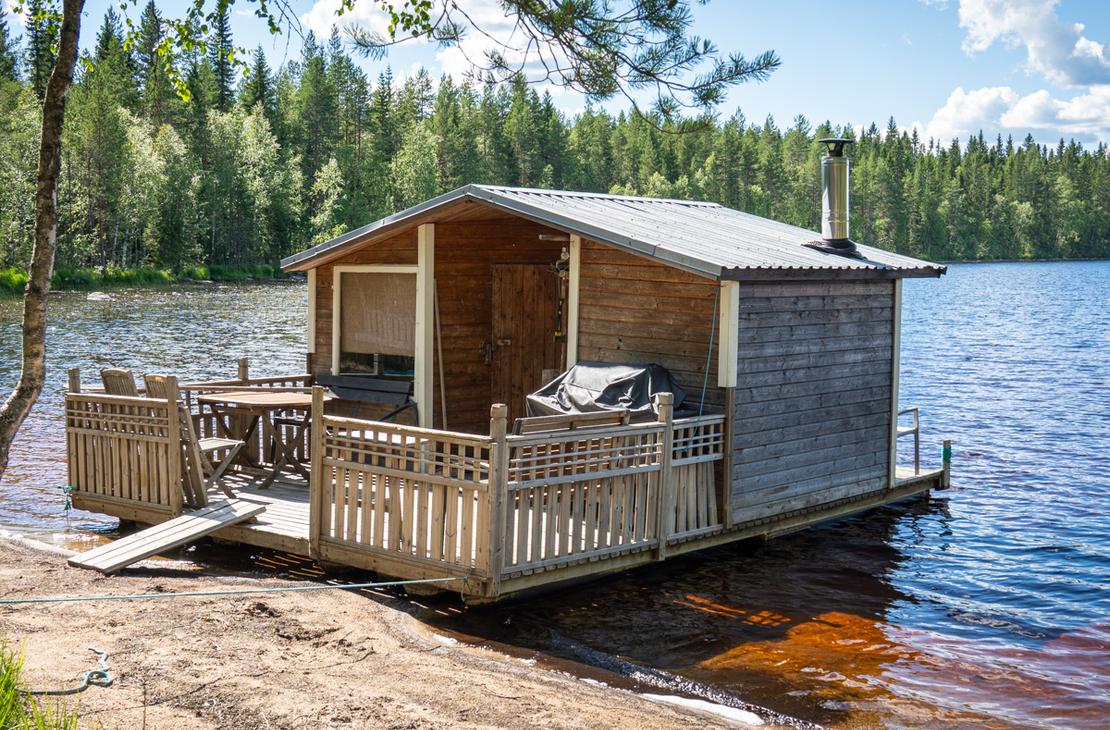 Puurakenteinen saunalautta ankkuroituna hiekkaiselle rannalle tyynen järven äärellä. Lautalla on terassi, jossa on pöytäryhmä ja grilli, sekä korkea metallinen piippu.