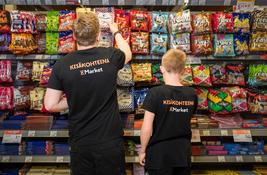 Kaksi mustiin t-paitoihin pukeutunutta henkil&ouml;&auml; selin kuvattuna K-Market Ilomantsin karkkihyllyn edess&auml;. Paitojen sel&auml;ss&auml; on teksti &rdquo;KES&Auml;KOHTEENA K-Market&rdquo;. Vasemmanpuoleinen henkil&ouml; kurkottaa ottamaan karkkipussia ylemm&auml;lt&auml; hyllylt&auml;, joka on t&auml;ynn&auml; v&auml;rikk&auml;it&auml; makeispusseja, kuten TV Mix, Marianne ja Dumle.