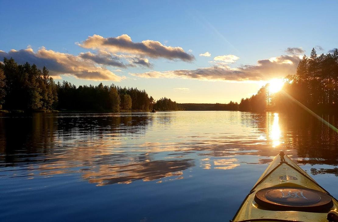 Kuva on otettu kajakista tai kanootista, ja se esittää tyyntä järveä auringonlaskun aikaan. Kajakin keula (vaaleankeltainen) on etualalla. Aurinko laskee tai nousee suoraan edessä, luoden kirkkaan heijastuksen veteen, joka on sinistä ja heijastaa taivaan pilviä. Järveä reunustaa tumma, tiheä metsä molemmin puolin. Kuva liittyy todennäköisesti Äksyt Ämmät -yrityksen melontapalveluihin.