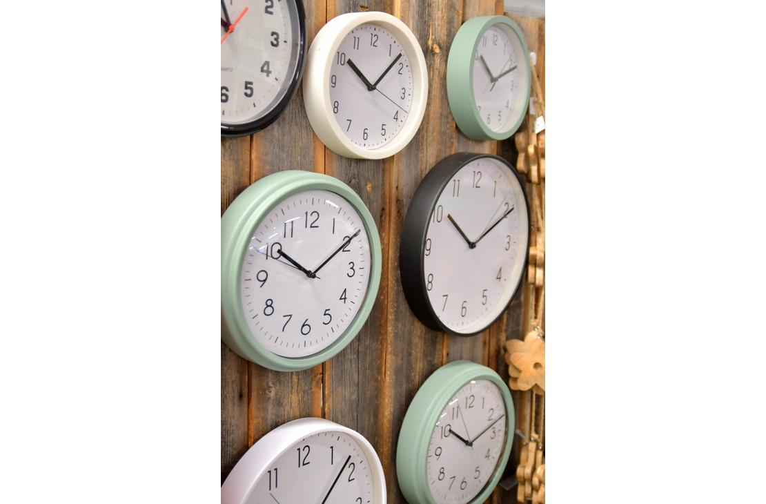 Ryhmä pyöreitä seinäkelloja, joiden kehykset ovat eri värisiä, kuten mintunvihreitä, valkoisia ja mustia, kiinnitettynä rouheaan puuseinään.