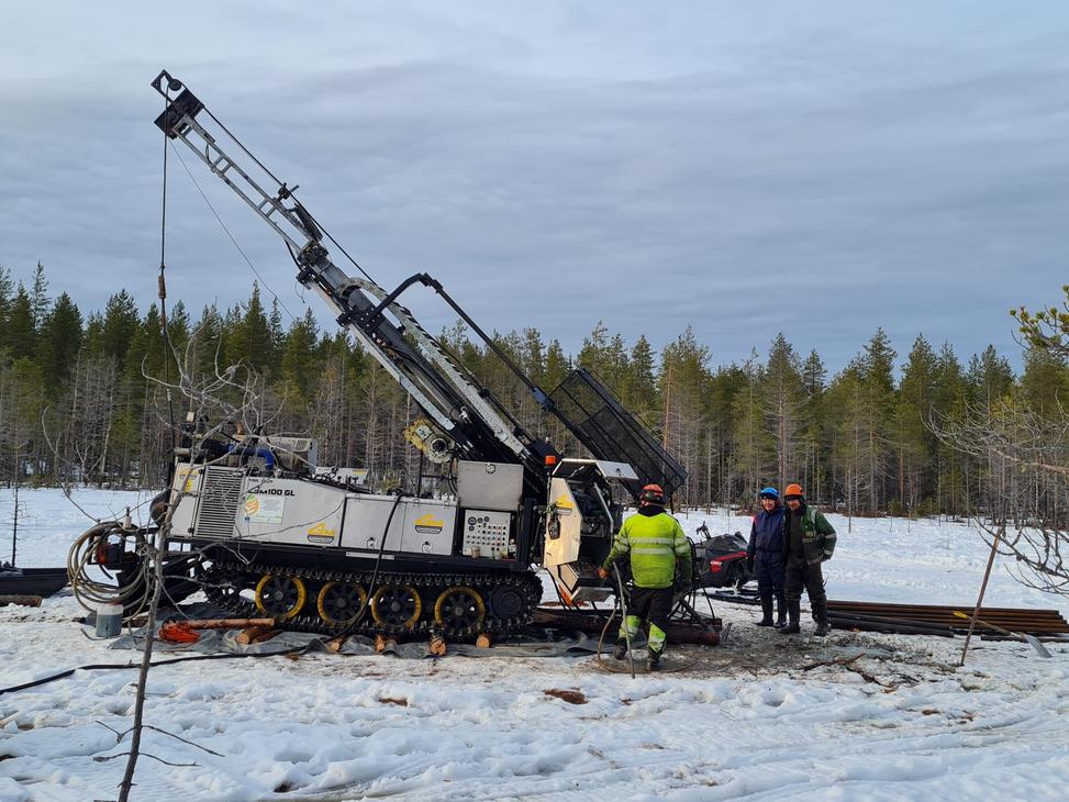 Kairaaja ja kaksi muuta henkilöä seisovat suuren telaketjuilla kulkevan kairauskoneen vieressä lumisella suolla. Taustalla on havumetsää ja pilvinen taivas.