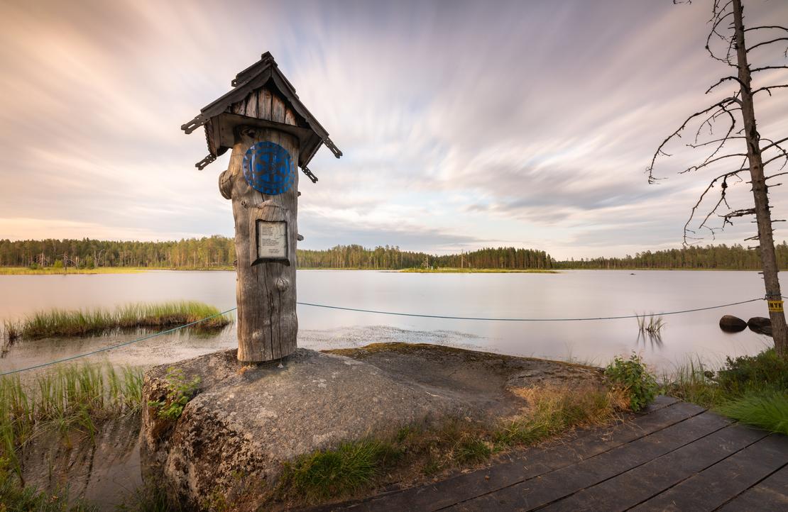 Kuva Suomen it&auml;isimm&auml;lt&auml; pisteelt&auml; auringonlaskun tai -nousun aikaan. Etualalla on puinen, veistetty muistomerkki tai pylv&auml;s, jonka p&auml;&auml;ll&auml; on pieni katos ja sininen, py&ouml;re&auml; symboli. Muistomerkki seisoo kivetyll&auml; alustalla veden &auml;&auml;rell&auml;. Taustalla avautuu laaja j&auml;rvimaisema, jonka vastarannalla on tummaa mets&auml;&auml;. Taivas on dramaattinen, pilvinen, ja siin&auml; n&auml;kyy pitk&auml;n valotusajan aiheuttamaa liikett&auml;. Oikealla n&auml;kyy kuollut, karu puu ja osittain puinen k&auml;velysilta.