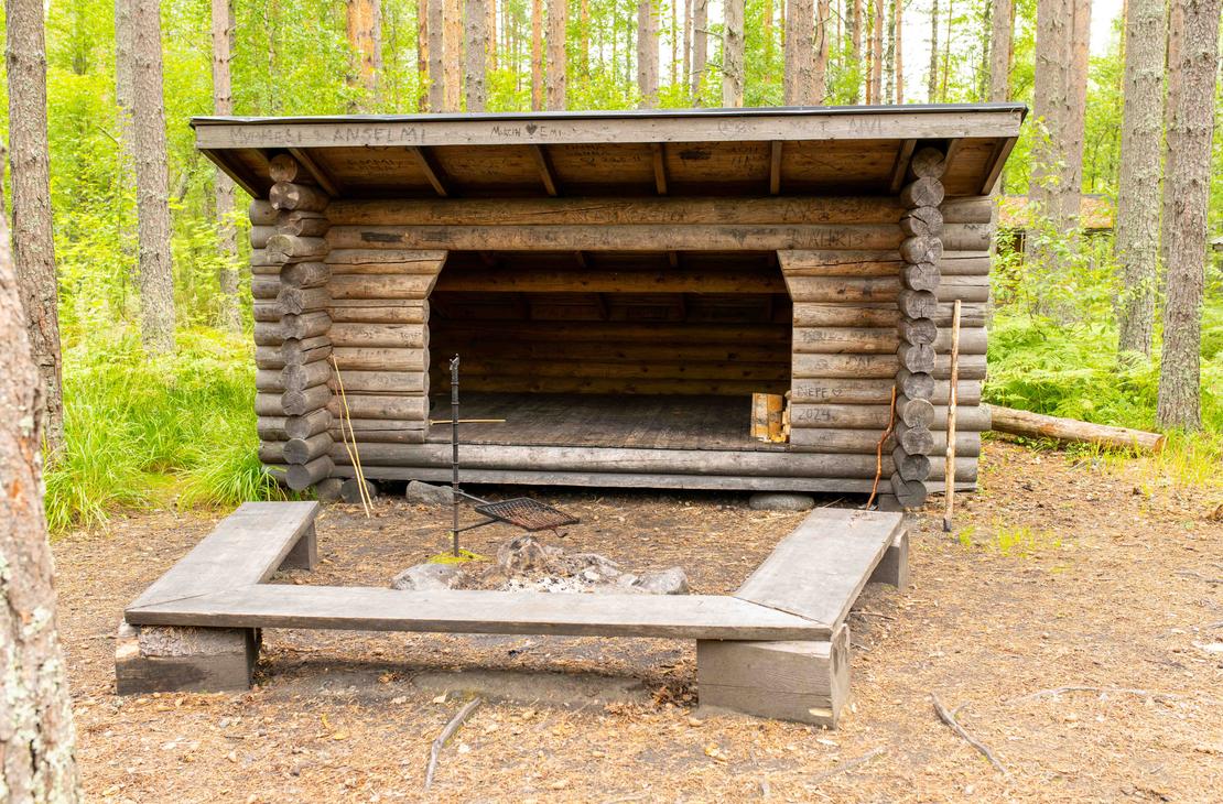Kookas hirsirakenteinen laavu, jonka edustalla on kivestä tehty nuotiopaikka, grilliritilä ja puupenkit. Laavun hirsipintoihin on kaiverrettu nimikirjoituksia.