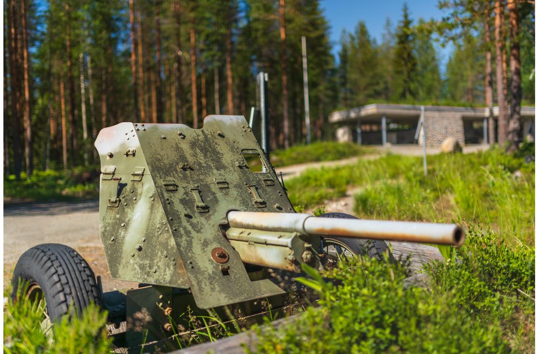 Kuva &Ouml;ykk&ouml;senvaaran taistelualueen muistomerkist&auml; tai n&auml;yttelypaikasta. Etualalla on ruosteinen, maastokuvioitu vanha tykki tai panssarintorjuntatykki. Tykki on esill&auml; mets&auml;isess&auml; ymp&auml;rist&ouml;ss&auml;, ja taustalla n&auml;kyy ep&auml;tarkkana matala, moderni, harmaasta betonista tehty rakennus, todenn&auml;k&ouml;isesti taistelualueen museo tai opastuskeskus.
