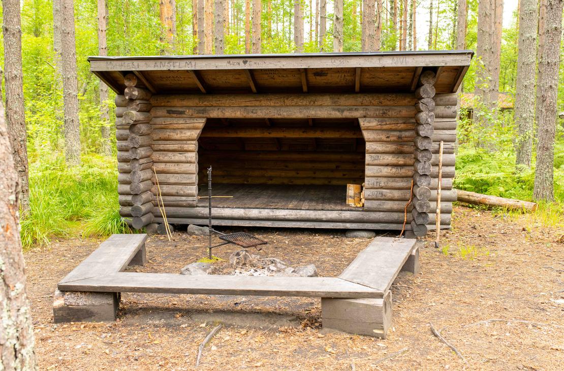 Kookas hirsirakenteinen laavu, jonka edustalla on kivestä tehty nuotiopaikka, grilliritilä ja puupenkit. Laavun hirsipintoihin on kaiverrettu nimikirjoituksia.