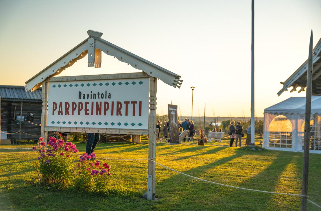 Lähikuva puisesta "Ravintola Parppeinpirtti" -kyltistä ilta-auringon paisteessa. Etualalla on kukkia, ja taustalla näkyy ihmisiä ravintolan pihapiirissä.