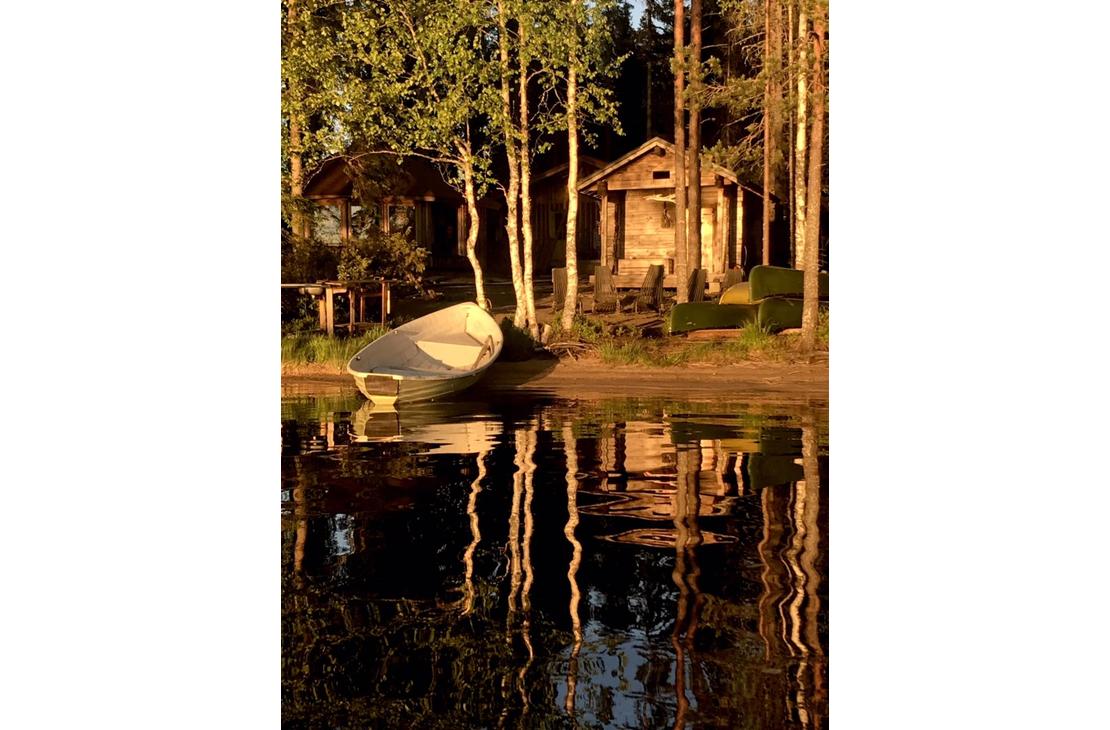 Kuva esittää Kotakylän rantasaunaa tai -mökkialuetta auringonlaskun aikaan. Etualalla on tummaa, tyyntä vettä, johon rannan puut ja rakennukset heijastuvat pitkinä varjoina. Rannalla, hiekalla tai nurmella, seisoo vaalea soutuvene kallellaan. Taustalla pystyssä olevien puunrunkojen takana näkyy kaksi pientä puurakennusta (mökit tai aitat), joita valaisee matala, lämmin auringonvalo.