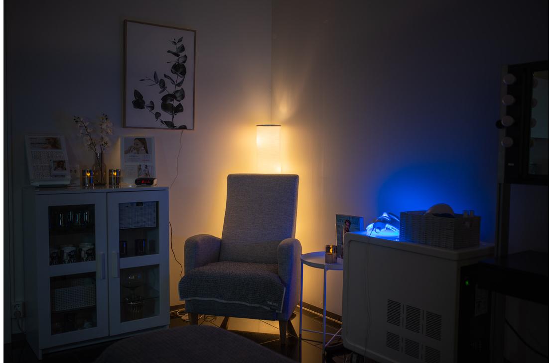 Kuva esittää Apteekkihoitola Alku -hoitolan tai vastaanoton huonetta, joka on valaistu tunnelmallisesti. Tilan keskellä on mukava, vaalea nojatuoli, jonka taakse sijoitettu valaisin antaa lämmintä, keltaista valoa. Oikealla puolella on hoitolaite tai tarjoiluvaunu, jonka sininen valo luo kontrastia. Vasemmalla on valkoinen kaappi ja seinällä kehystetty taulu. Tila on rauhallinen ja hämärä.