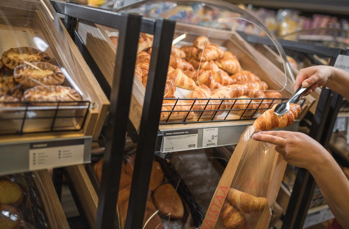 Kädet siirtävät metallisilla ottimilla voicroissantia paistopisteen lokerosta ikkunalliseen paperipussiin K-Market Ilomantsissa. Lokeron alla olevassa tuoteselosteessa lukee ”PIRKKA VOICROISSANT 50G”. Vasemmalla puolella olevassa lokerossa on esillä karjalanpiirakoita.