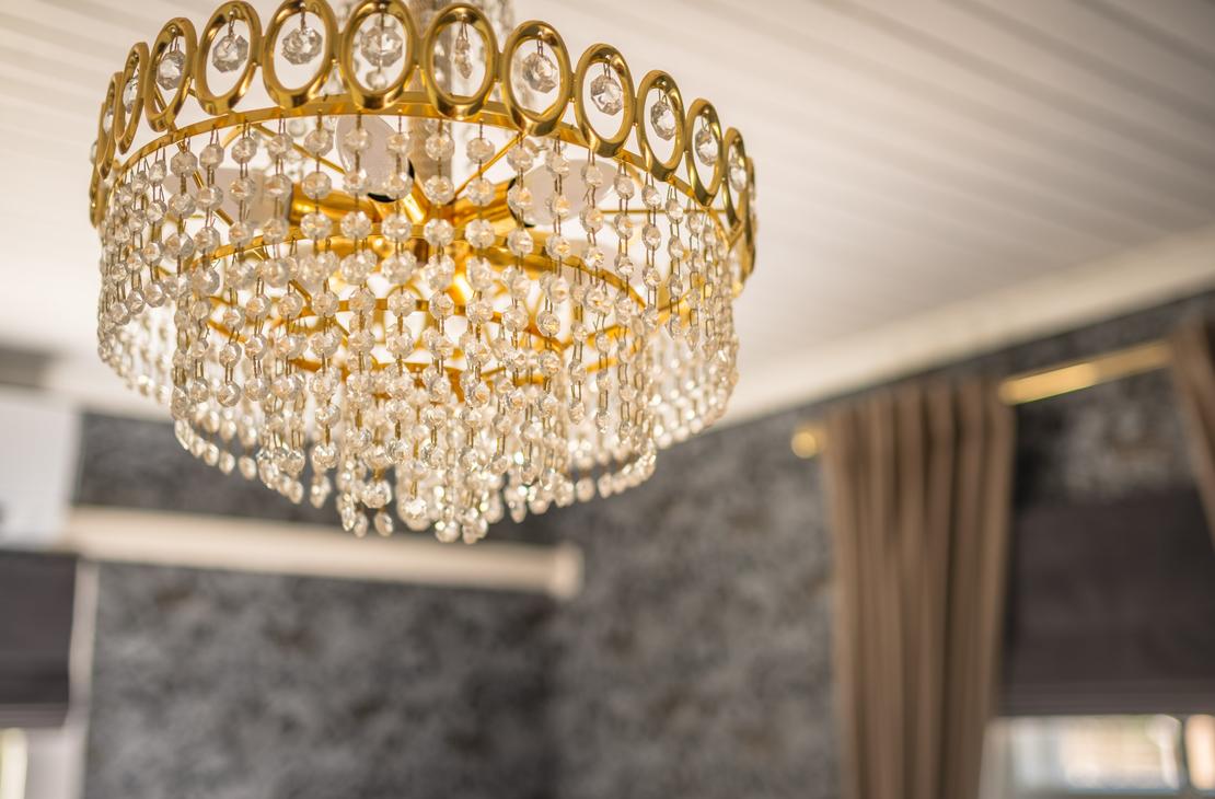 Lähikuva kristallikruunusta Matkailukeskus Puu-Juuka Oy:n sisätiloissa. Kruunu on suuri, kultaisen värinen ja siinä on useita kerroksia roikkuvia kristalleja. Kruunu roikkuu valkoisesta, lankulla vuoratusta katosta. Tausta on epätarkka, mutta siellä näkyy tummanharmaata tai mustaa seinämateriaalia ja ruskeita verhoja.