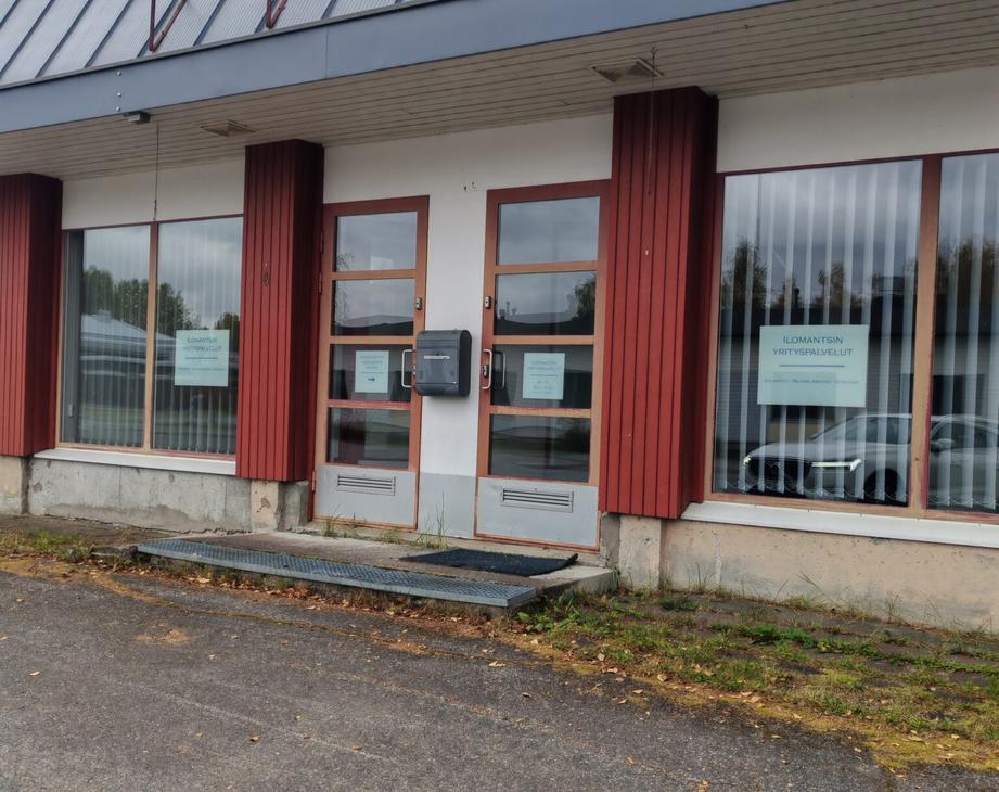 Ilomantsin Yrityspalvelut -toimiston sisäänkäynti ulkoapäin. Yksikerroksisen rakennuksen julkisivussa on valkoista seinää, pystysuuntaisia punaruskeita puupaneeleita sekä suuria ikkunoita pystylamellikaihtimilla. Keskellä on kaksi ruskeakehyksistä lasiovea, joiden välissä on harmaa postilaatikko ja edessä metalliritiläinen askelma mattoineen. Oikeanpuoleisessa ikkunassa on valkopohjainen kyltti, jossa lukee "ILOMANTSIN YRITYSPALVELUT KIRJANPITO - PALKANLASKENTA - VEROASIAT". Etualalla on asfaltoitu piha-alue, ja rakennuksen seinustalla kasvaa hieman ruohoa.