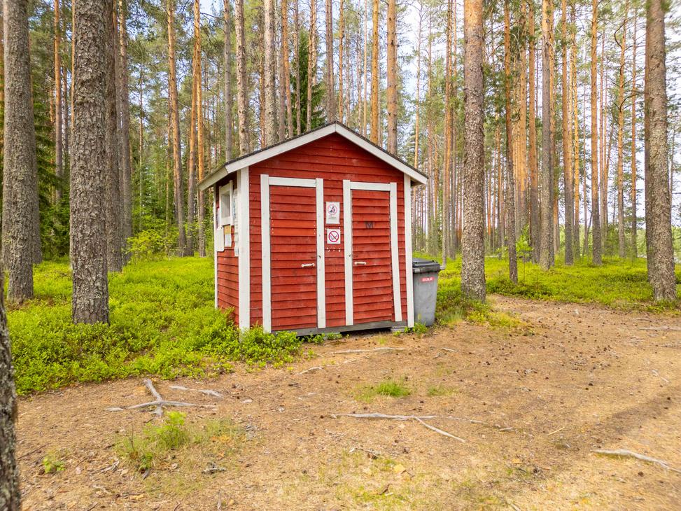 Punainen valkonurkkainen kuivakäymälä rakennus mäntymetsän keskellä. Rakennuksen vieressä on harmaa roska-astia ja maasto on käpystä kangasmaata.