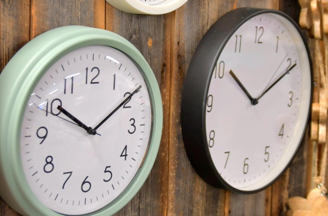 Ryhmä pyöreitä seinäkelloja, joiden kehykset ovat eri värisiä, kuten mintunvihreitä, valkoisia ja mustia, kiinnitettynä rouheaan puuseinään.