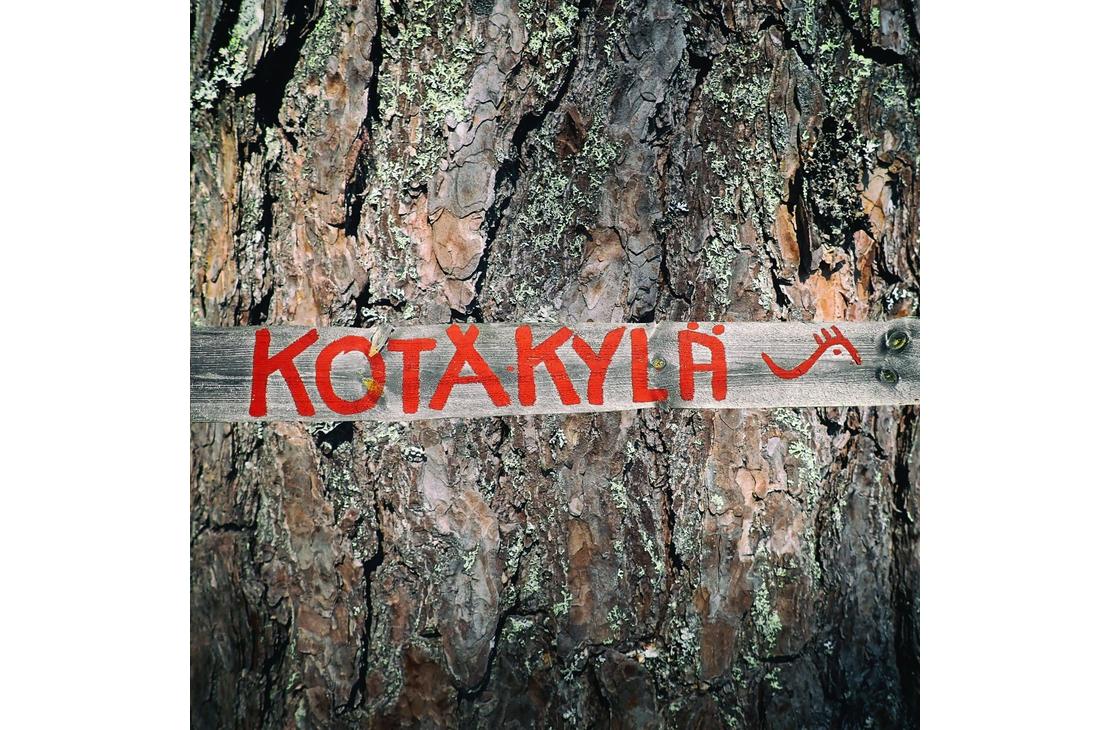 Lähikuva vanhan puun (todennäköisesti männyn) karkeasta, halkeilevasta kuoresta. Puunrunkoa vasten on kiinnitetty vaakasuora, vaaleampi puulauta, johon on maalattu kirkkaan punaisella tekstillä sana "KOTAKYLÄ". Tekstin oikeassa reunassa on pieni, punainen, tyylitelty kuvio, joka muistuttaa sarvipäätä tai eläintä.
