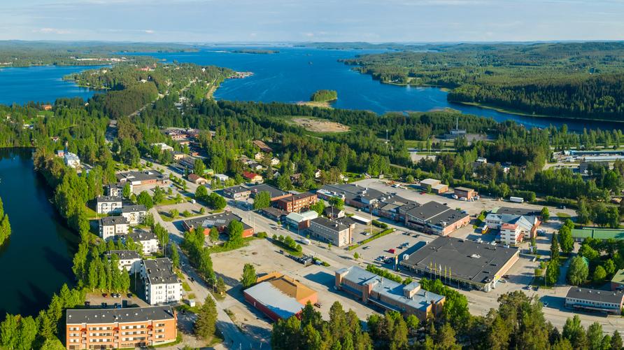 Laaja ilmakuva taajamasta, joka sijaitsee vehreiden metsien ja laajan, sinisen järvimaiseman keskellä aurinkoisena päivänä. Alueella näkyy asuin- ja liikerakennuksia.