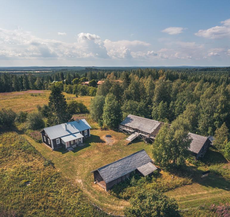 Ilmakuva Makkolan museotilasta, joka koostuu useista perinteisistä, tummista hirsirakennuksista harmailla katoilla. Rakennukset sijaitsevat vihreällä niityllä, jota ympäröi tiheä metsä ja aukea pelto. Piha-alueella näkyy ajopolkuja ja pieni avoin alue rakennusten välissä.