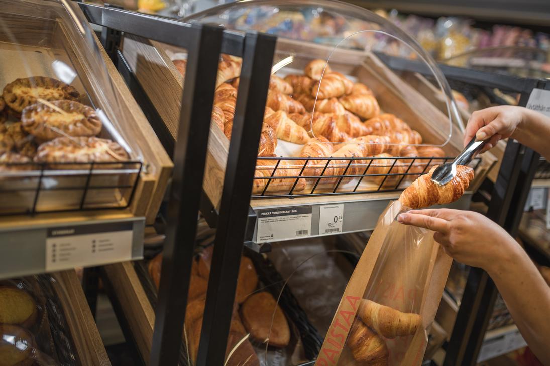 K&auml;det siirt&auml;v&auml;t metallisilla ottimilla voicroissantia paistopisteen lokerosta ikkunalliseen paperipussiin K-Market Ilomantsissa. Lokeron alla olevassa tuoteselosteessa lukee &rdquo;PIRKKA VOICROISSANT 50G&rdquo;. Vasemmalla puolella olevassa lokerossa on esill&auml; karjalanpiirakoita.