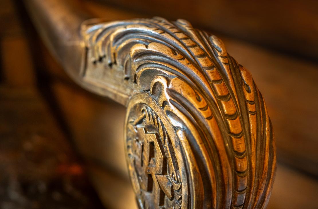 Lähikuva tumman puisen kaiteen tai tuolin käsinojan päädystä, jossa on taidokkaasti veistetty, koristeellinen kuvio. Kaiverrus on pyöreä ja kupera, muistuttaen simpukankuorta tai sulkakuviointia, ja sen keskellä on erottuva, geometrinen kuvio. Käsinoja on kiillotettu ja kiinnittyy tummaan pylvääseen. Tausta on epätarkka, mutta siinä näkyy puupintoja.