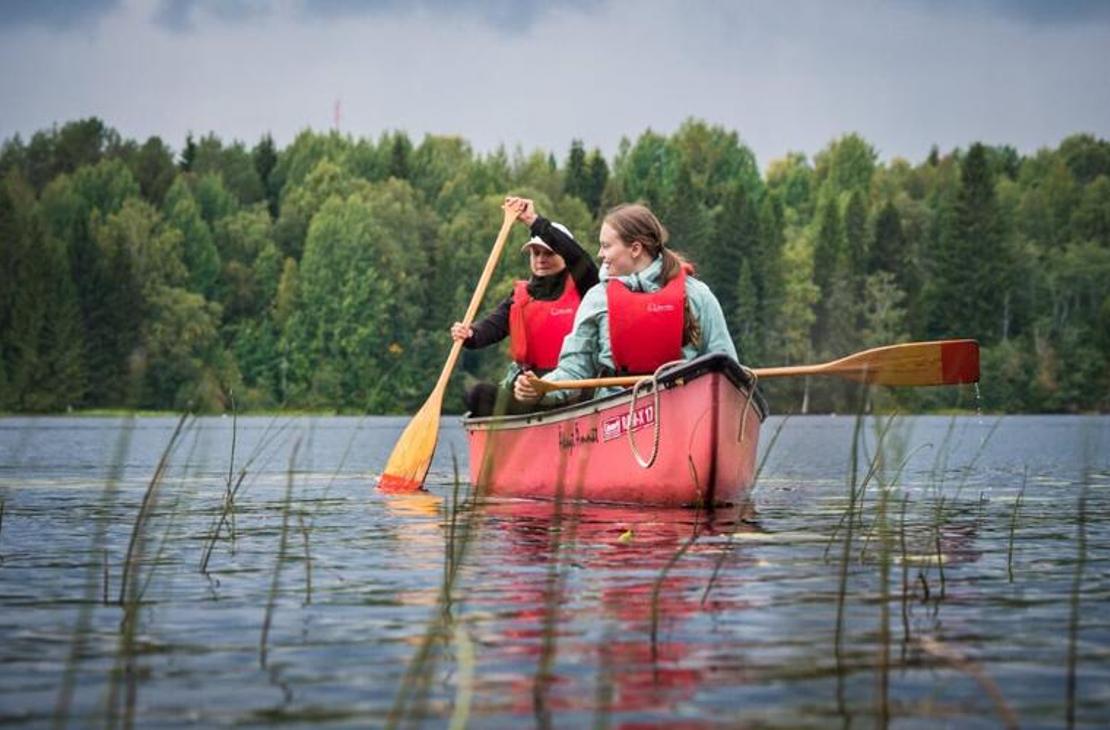 Kaksi henkilöä, nainen etualalla ja mies takana, melovat punaisella avokanootilla tyynellä järvellä. Molemmilla on yllään kirkkaan punaiset pelastusliivit. He pitelevät puisia meloja, joissa on keltaiset lavat. Etualalla vedestä nousee pitkiä, ohuita ruohoja. Taustalla on tiheä, vihreä metsä. Kuva liittyy todennäköisesti Äksyt Ämmät -yrityksen melontapalveluihin.
