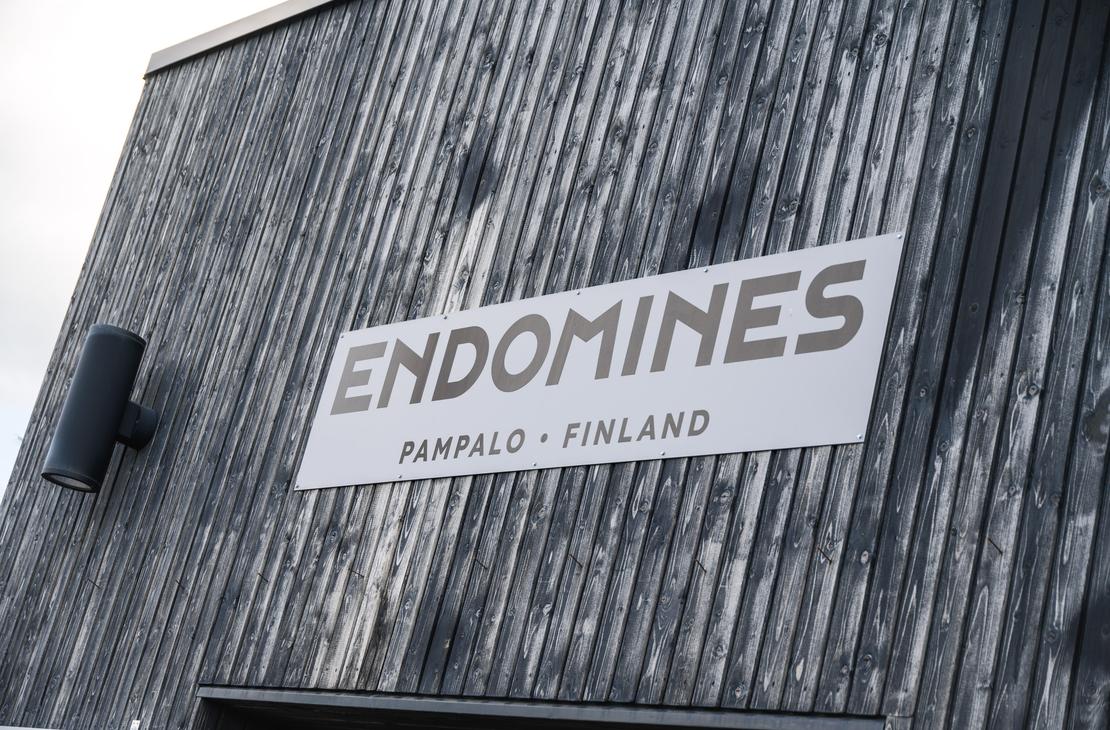 Harmaa pystysuuntainen puupaneeliseinä, johon on kiinnitetty valkoinen suorakaiteen muotoinen kyltti. Kyltissä lukee suurin kirjaimin "ENDOMINES" ja sen alapuolella pienemmällä "PAMPALO • FINLAND". Seinän vasemmassa reunassa on musta lieriömäinen ulkovalaisin.