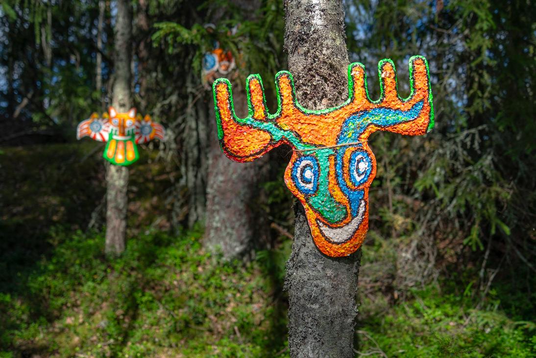 Puiden runkoihin kiinnitettyjä värikkäitä, primitiivisiä naamioita ja eläinhahmoja, kuten oranssi-vihreä hirvi- tai peura-aiheinen taideteos.