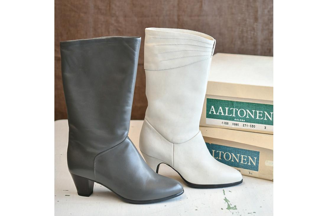 Harmaa ja valkoinen siro nahkasaapas aseteltuna vierekkäin. Taustalla näkyy Aaltonen-kenkälaatikoita.