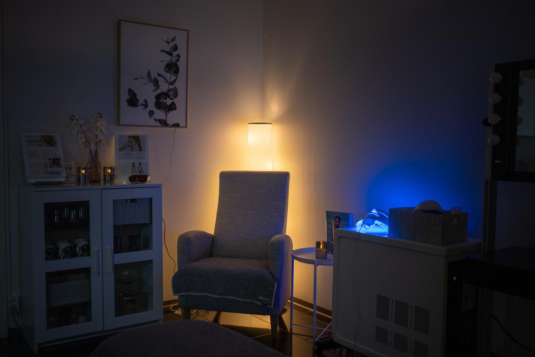 Kuva esittää Apteekkihoitola Alku -hoitolan tai vastaanoton huonetta, joka on valaistu tunnelmallisesti. Tilan keskellä on mukava, vaalea nojatuoli, jonka taakse sijoitettu valaisin antaa lämmintä, keltaista valoa. Oikealla puolella on hoitolaite tai tarjoiluvaunu, jonka sininen valo luo kontrastia. Vasemmalla on valkoinen kaappi ja seinällä kehystetty taulu. Tila on rauhallinen ja hämärä.