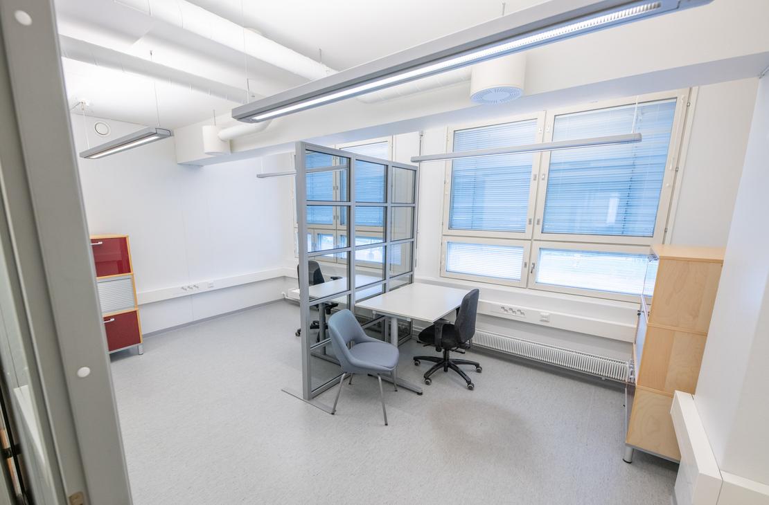 Kuva esittää Liikekulman Toimistohotellin sisätiloja. Käytävä on valoisa ja moderni, jossa on vaalea lattia ja valkoiset seinät. Oikealla on pitkä käytävä, jossa on oviaukkoja ja työpöytiä. Vasemmalla on pyöreitä mustia pöytiä ja tuoleja. Keskellä näkyy kirkkaan limevihreä ovi, jossa on logo, ja taaempana oranssin ja keltaisen sävyisiä ovia, jotka viittaavat erilaisiin toimistoihin.