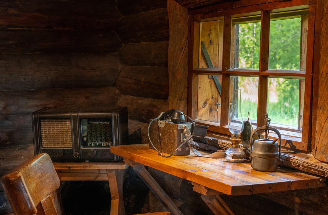 Kuva Rajakenraalin majan sis&auml;tilasta, jossa esitet&auml;&auml;n perinteist&auml; hirsist&auml; sisustusta. L&auml;hikuva keskittyy puiseen ikkunanp&ouml;yt&auml;&auml;n, jolla on vanhanaikainen termospullo ja metallinen astia. P&ouml;yt&auml; on sijoitettu ikkunan eteen, josta siivil&ouml;ityy sis&auml;&auml;n valoa ja n&auml;kyy ulkoa vihre&auml;&auml; kasvillisuutta. Vasemmalla tumman hirsisein&auml;n vieress&auml; n&auml;kyy vanha p&ouml;yt&auml;radio.