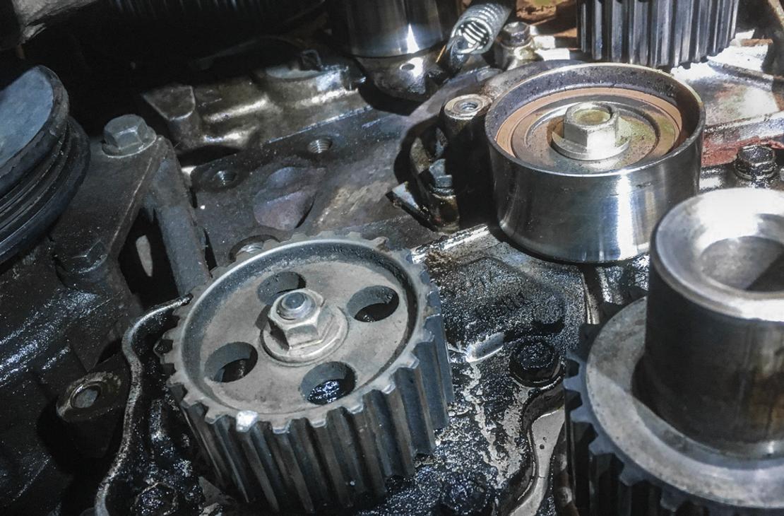 Lähikuva autonmoottorin osista, todennäköisesti jakopään alueelta. Kuvassa näkyy kaksi hammaspyörää ja yksi kiristinrulla tai vastaava metallinen pyörä. Osat ovat tummat, kiiltävät ja mahdollisesti öljyiset. Kohtaus on otettu moottoritilassa korjauksen tai huollon aikana.