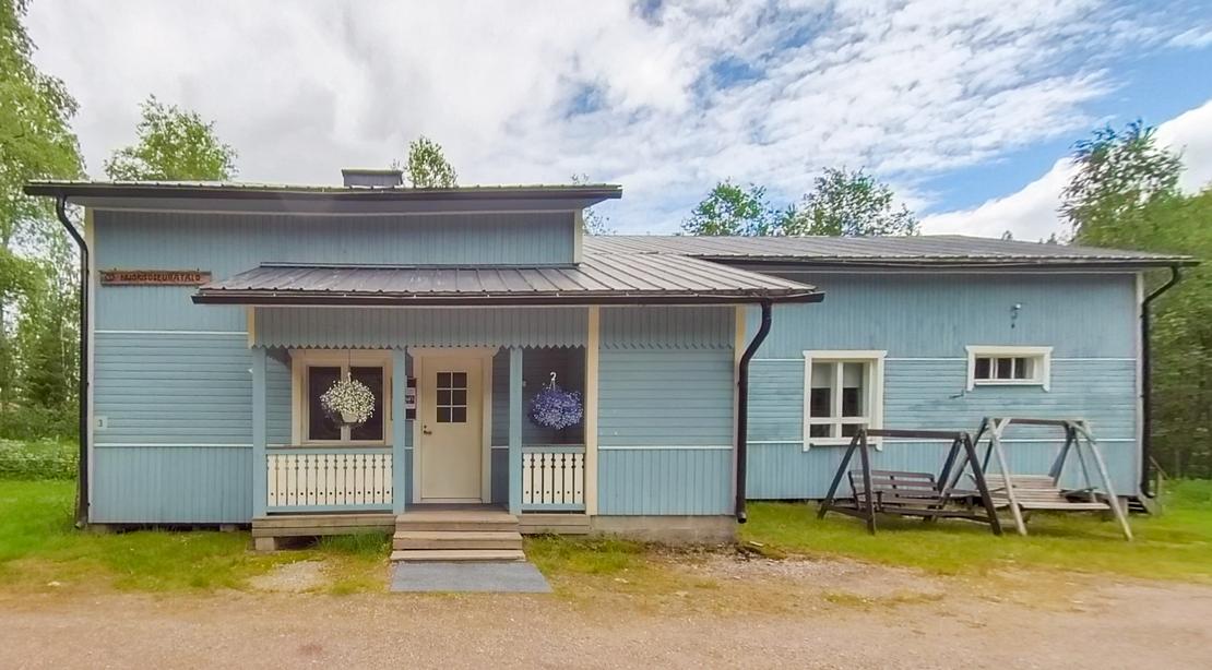 Kuva esittää matalaa, modernia, vaaleansinistä puutaloa tai rakennusta, jossa on tumma katto. Talon keskellä on pieni, katettu kuisti, jonka edessä on portaat. Kuistin oven yläpuolella näkyy pyöreä, koristeellinen valaisin tai symboli. Talon edessä on soraa tai hiekkamaata, ja oikealla sivulla on tumma, metallinen keinu tai teline nurmikolla. Taustalla näkyy rehevää kasvillisuutta ja metsää.