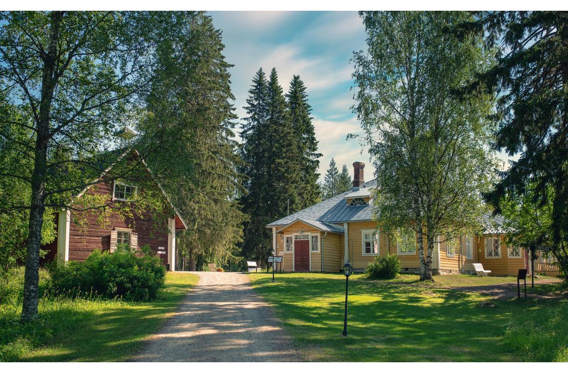 Kuva Möhkön ruukkimuseon alueelta esittää historiallista pihapiiriä. Oikealla on pitkä, matala, keltainen puutalo punaisella ovella ja harmaalla katolla. Vasemmalla näkyy korkeampi, punamultainen ulkorakennus tai aitta. Rakennusten välissä kulkee hiekkainen tie, ja niitä ympäröi vanhoja koivuja ja havupuita aurinkoisessa kesäympäristössä.