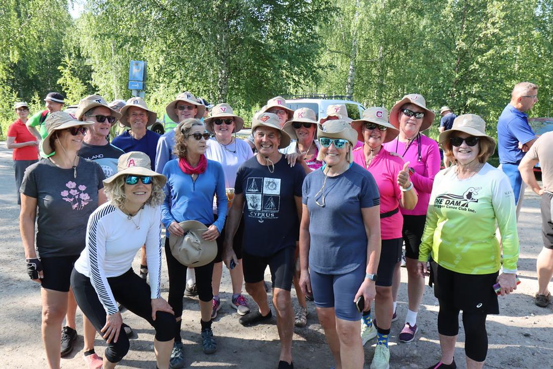 Ryhm&auml; aurinkohattuihin pukeutuneita hymyilevi&auml; retkeilij&ouml;it&auml; seisoo hiekkatiell&auml; puiden katveessa. Useimpien hatuissa on yksitt&auml;inen kirjainmerkki.