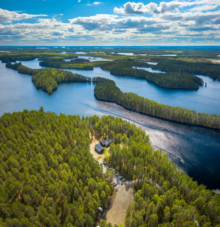 Ilmakuva Petkelj&auml;rvi Centerin alueesta ja Petkelj&auml;rven kansallispuiston maisemista. Etualalla on tihe&auml;&auml;, vihre&auml;&auml; mets&auml;&auml; ja avoin, hiekkainen alue, jossa n&auml;kyy muutama tummakattoinen rakennus (Petkelj&auml;rvi Center). Taustalla avautuu laaja j&auml;rvimaisema, jossa tummansininen vesi kietoutuu lukuisten vihreiden harjujen ja niemien ymp&auml;rille. Kirkas sininen taivas, jossa on valkoisia pilvi&auml;.