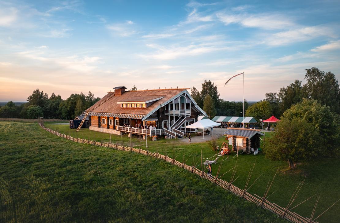 Viistosta yläkulmasta otettu kuva Parppeinpirtin hirsirakennuksesta ja sen pihasta. Etualalla on perinteinen riukuaita, joka reunustaa vihreää niittyä. Taivaanrannassa näkyy auringonlaskun värejä.