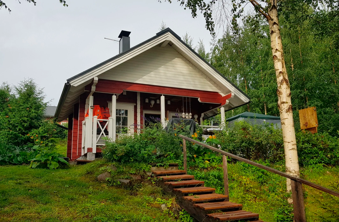 Kuva esittää Tujanranta B&B:n pientä, perinteistä punamultamaalilla maalattua mökkiä tai aittarakennusta. Rakennuksessa on valkoiset ikkunankarmit ja päätykolmio sekä tumma harjakatto. Mökin edessä on rinteessä puiset portaat ja kaiteet. Rakennusta ympäröi runsas, vihreä kasvillisuus ja koivuja.