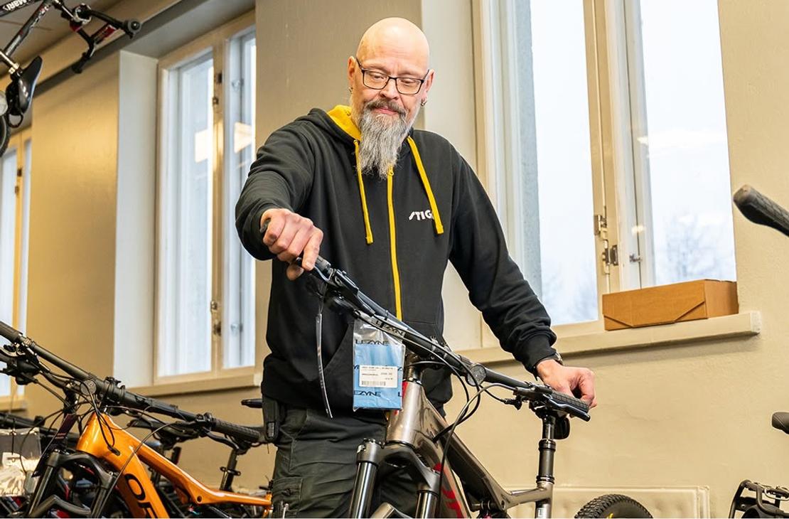 Silmälasipäinen, parrakas mies tarkastelemassa polkupyörää myymälässä. Kuvan alalaidassa on Lieksan Koneasema -tekstilogo.